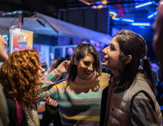 Three friends laughing and dancing under blue neon lights at a crowded indoor nightlife venue; center person in a pastel-striped sweater, curly-haired friend on the left, and a smiling person in a light puffer vest on the right.