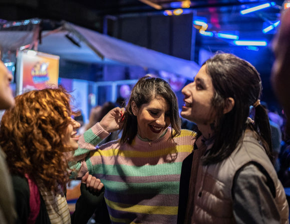 Three friends laughing and dancing under blue neon lights at a crowded indoor nightlife venue; center person in a pastel-striped sweater, curly-haired friend on the left, and a smiling person in a light puffer vest on the right.