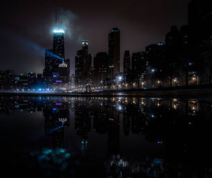 Moody nighttime city skyline with illuminated skyscrapers and a blue searchlight, mist drifting above calm water and crisp reflections of streetlights along a waterfront promenade.