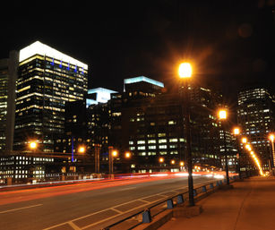 Nighttime downtown skyline with illuminated office skyscrapers, a lamp-lit bridge in the foreground and red and white traffic light trails from long-exposure cars.