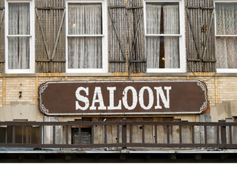 Rustic Old West-style "SALOON" sign on a weathered brick facade with a wooden balcony, shuttered second-floor windows and lace curtains.