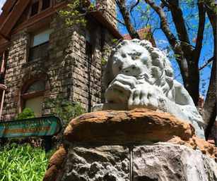 Regal stone lion sculpture lounging on a sandstone pedestal outside a historic stone mansion, framed by leafy trees and a bright blue sky.