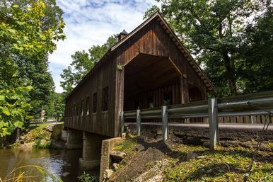 Rustic wooden covered bridge spanning a shallow creek in a forested rural setting, sunlit summer sky and roadside guardrail in the foreground