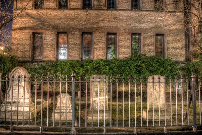 Historic churchyard with weathered stone headstones behind a wrought-iron fence, set against a lit brick building with leaded windows and ivy — nighttime urban cemetery scene.