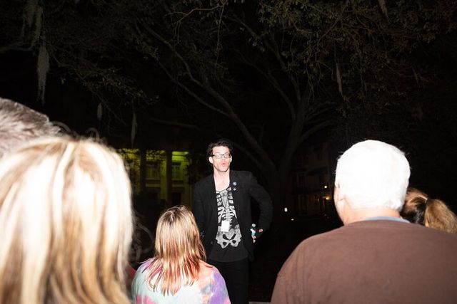 Nighttime outdoor tour guide in a skeleton shirt speaking to a small group under a large oak tree with Spanish moss and a lit building facade in the background