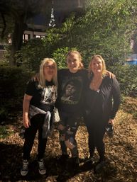 Three friends smiling and posing at night in a leafy park, arms around each other; center wearing a The Exorcist graphic tee and ripped jeans, others in black outfits, warm outdoor lighting with fallen leaves underfoot and a lit spire visible in the background.