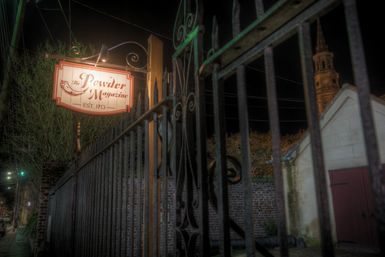 Moody nighttime shot of an illuminated vintage powder-magazine sign (est. 1713) hanging above an ornate iron fence with a brick wall and distant church steeple.