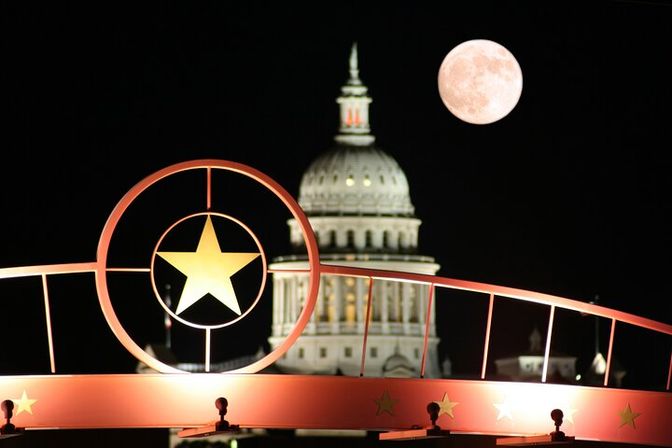 Glowing red metal star arch in the foreground framing an illuminated state capitol dome and a bright full moon against a black night sky.