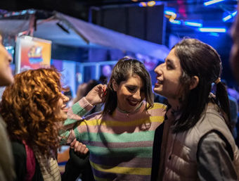 Three friends laughing and dancing under neon lights at a lively bar or nightclub during a city night out; woman in a pastel striped sweater smiles between two friends.