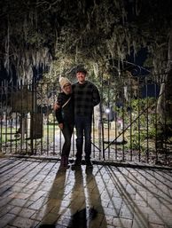 Smiling couple in winter coats posing at night by an ornate iron gate beneath Spanish moss-draped oak trees, long shadows on a wet brick path in a historic cemetery.