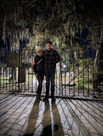 Smiling couple in winter coats posing at night by an ornate iron gate beneath Spanish moss-draped oak trees, long shadows on a wet brick path in a historic cemetery.
