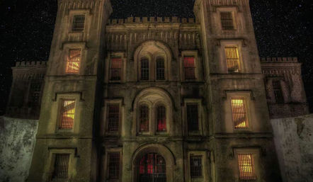 Eerie historic stone facade with towers and barred, warmly lit windows glowing against a starry night sky