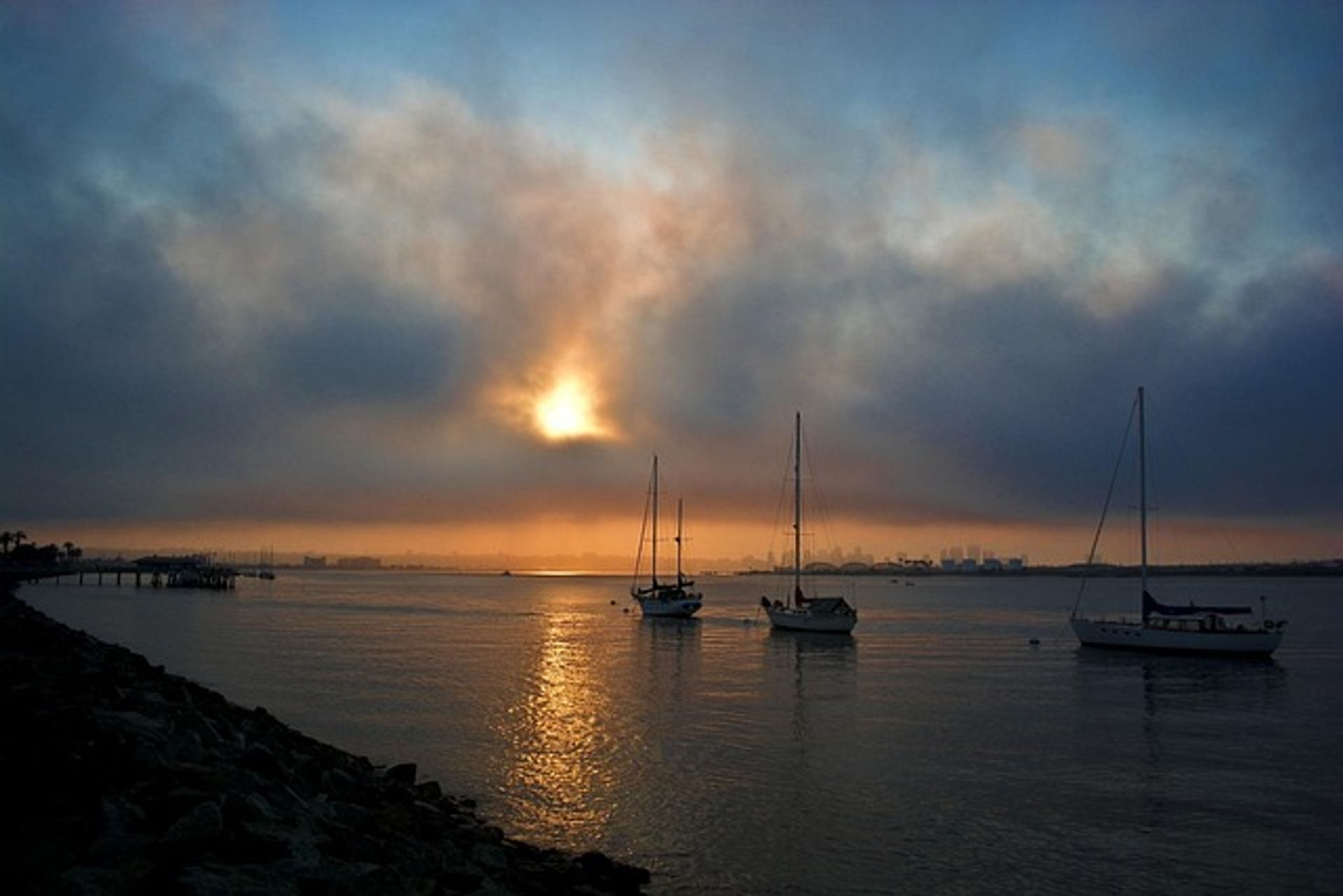 Misty coastal harbor at sunset with a golden sun reflecting on calm water, three anchored sailboats and a rocky shoreline with a pier