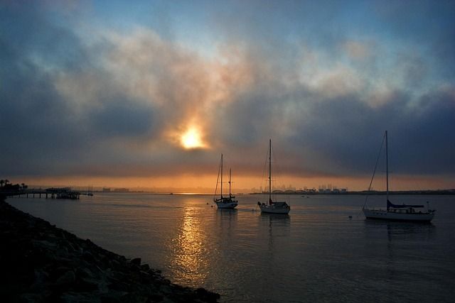 Misty coastal harbor at sunset with a golden sun reflecting on calm water, three anchored sailboats and a rocky shoreline with a pier