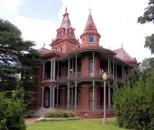 Red-brick Victorian mansion with conical turrets and decorative spires, ornate cast-iron wraparound porch and tall columns, set behind a manicured lawn and mature trees.