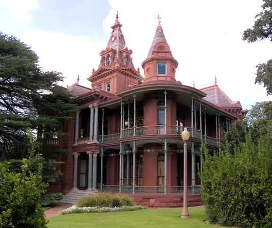 Red-brick Victorian mansion with conical turrets and decorative spires, ornate cast-iron wraparound porch and tall columns, set behind a manicured lawn and mature trees.