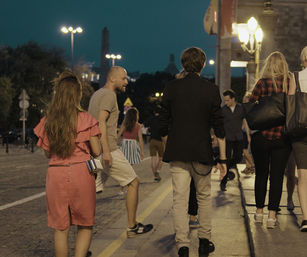 People strolling on a cobblestone downtown street at dusk, illuminated by lampposts and warm evening lights, casual summer attire.