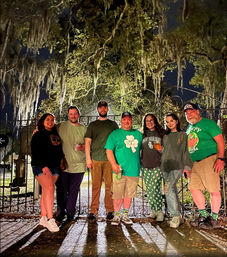Seven people in green St. Patrick's attire posing at night in front of a wrought-iron gate beneath Spanish-moss-draped live oaks, holding drinks and smiling