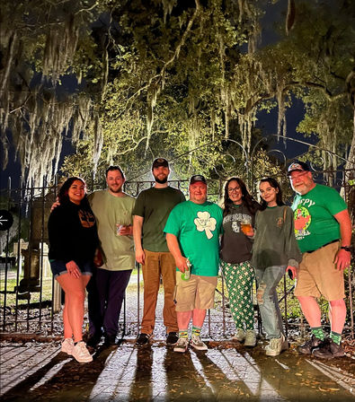 Seven people in green St. Patrick's attire posing at night in front of a wrought-iron gate beneath Spanish-moss-draped live oaks, holding drinks and smiling