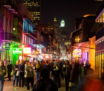 Nighttime New Orleans French Quarter street scene with crowds, colorful neon signs, wrought-iron balconies and lively bar and restaurant lights.
