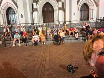 Group of people seated on wide stone steps of a Gothic-style church with arched wooden doors, gathered on a brick plaza at night while a person’s partial selfie appears in the foreground.
