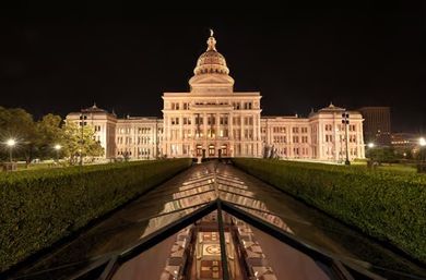 Night view of the illuminated Texas State Capitol in Austin, TX, grand domed building with symmetrical facade reflected in a glass-lined pool and flanked by manicured hedges