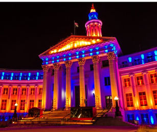 Neoclassical civic building with tall Corinthian columns and domed cupola, steps to the entrance, dramatically lit in vibrant purple, blue and orange at night