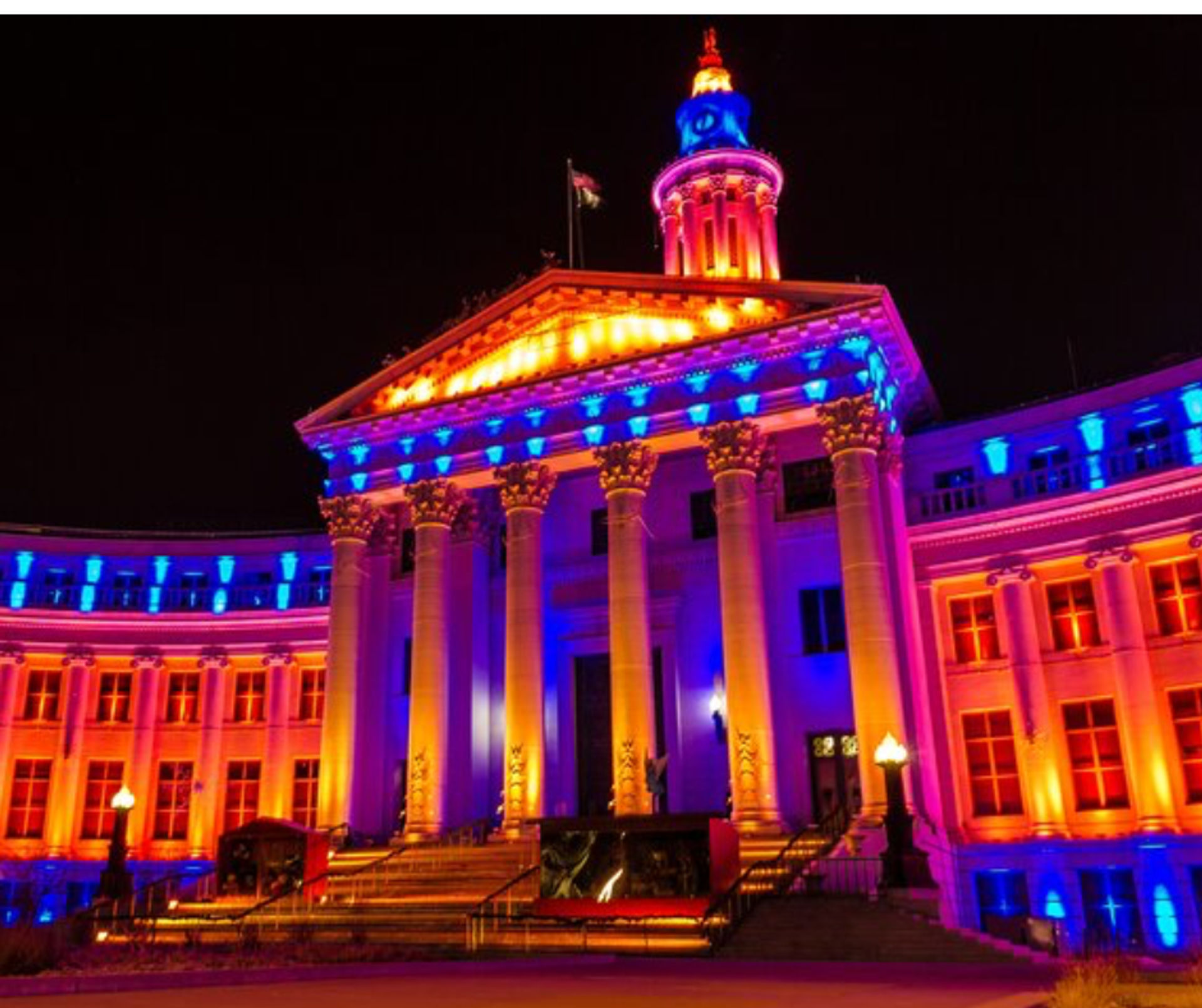 Neoclassical civic building at night — grand columned facade and cupola dramatically illuminated in vibrant blue, purple, and orange lights, a colorful nighttime landmark with steps and lampposts.