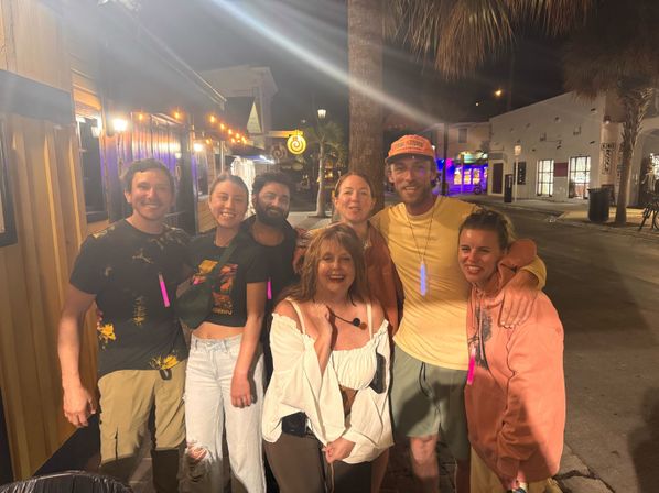Seven friends smiling for a nighttime photo on a palm-tree-lined downtown street under string lights and lit storefronts, wearing casual outfits and glow necklaces — fun night out vibe.
