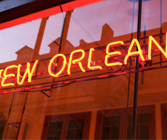 Vibrant red-orange neon sign reading "NEW ORLEANS" glowing in a storefront window with warm reflections of historic building facades.