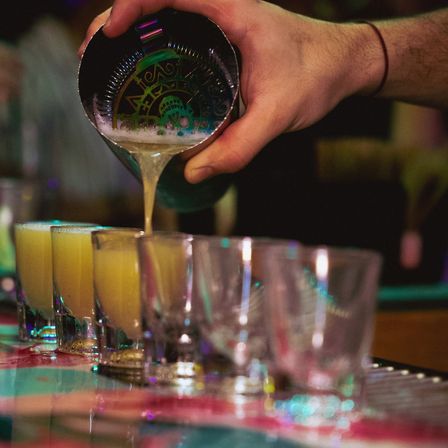 Bartender pouring frothy yellow cocktails from a shaker into lined shot glasses on a colorful bar counter, lively nightlife scene