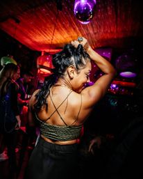 Woman dancing in a vibrant nightclub under colorful disco lights and hanging lamps, wearing a strappy crop top with braided hair and arms raised on a crowded dance floor