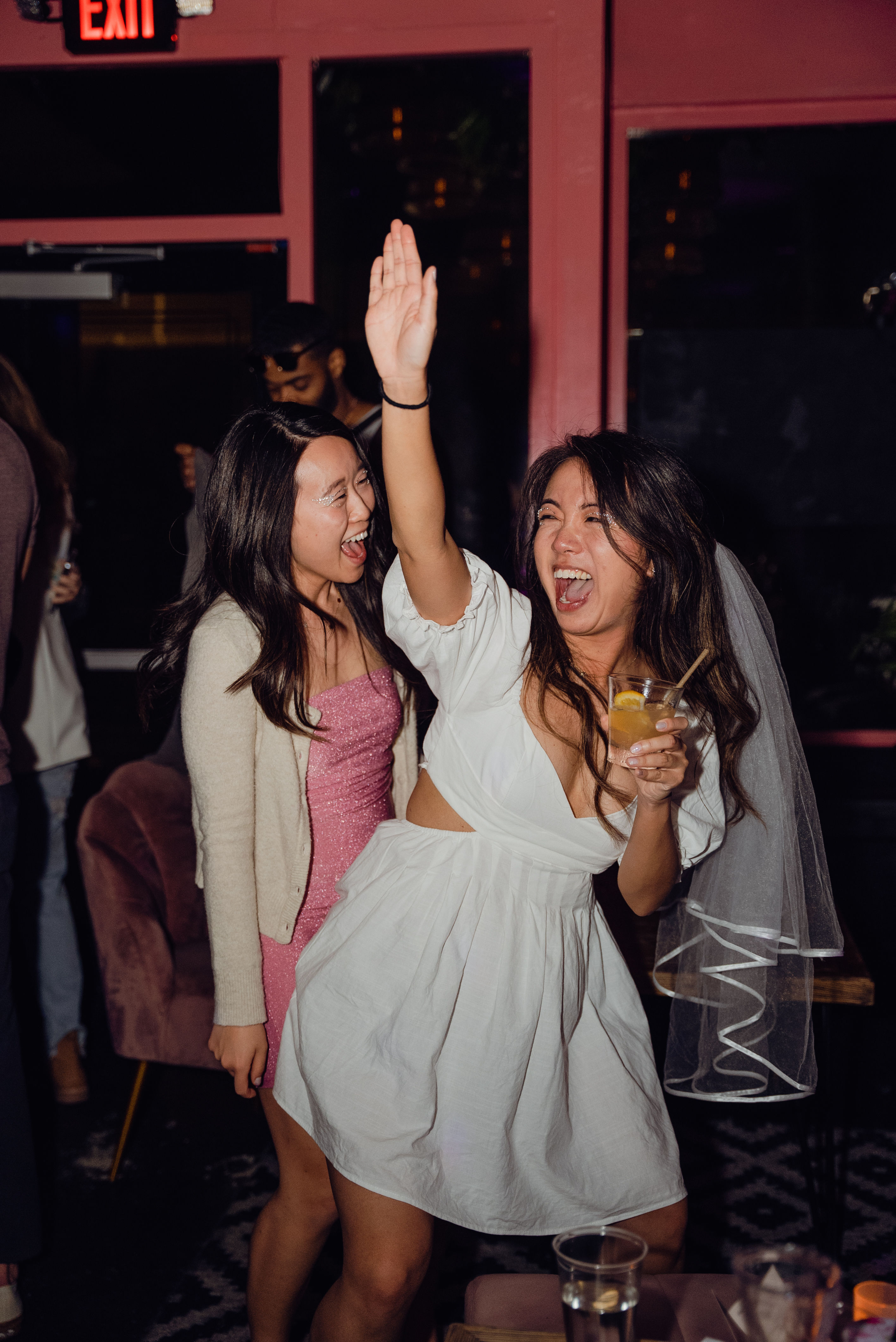 Cheerful bachelorette at a lively bar: woman in a white dress and veil raises her hand and holds a cocktail while a friend in a pink sparkly dress laughs nearby, nightlife celebration.