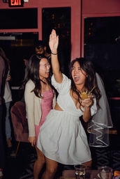 Cheerful bachelorette at a lively bar: woman in a white dress and veil raises her hand and holds a cocktail while a friend in a pink sparkly dress laughs nearby, nightlife celebration.