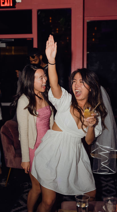 Cheerful bachelorette at a lively bar: woman in a white dress and veil raises her hand and holds a cocktail while a friend in a pink sparkly dress laughs nearby, nightlife celebration.