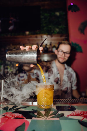 Bartender pouring bright orange tropical cocktail from a shaker into a sugar-rimmed glass as smoky vapor swirls over a colorful flamingo-patterned bar