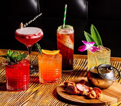 Vibrant craft cocktails on a wooden bar table: frothy red drink with a lavender sprig, mint-garnished ruby cocktail, orange citrus tumbler with rosemary, cucumber-studded highball with a striped straw, orchid-topped tropical cocktail, and a wooden board of charcuterie and crispy snacks.