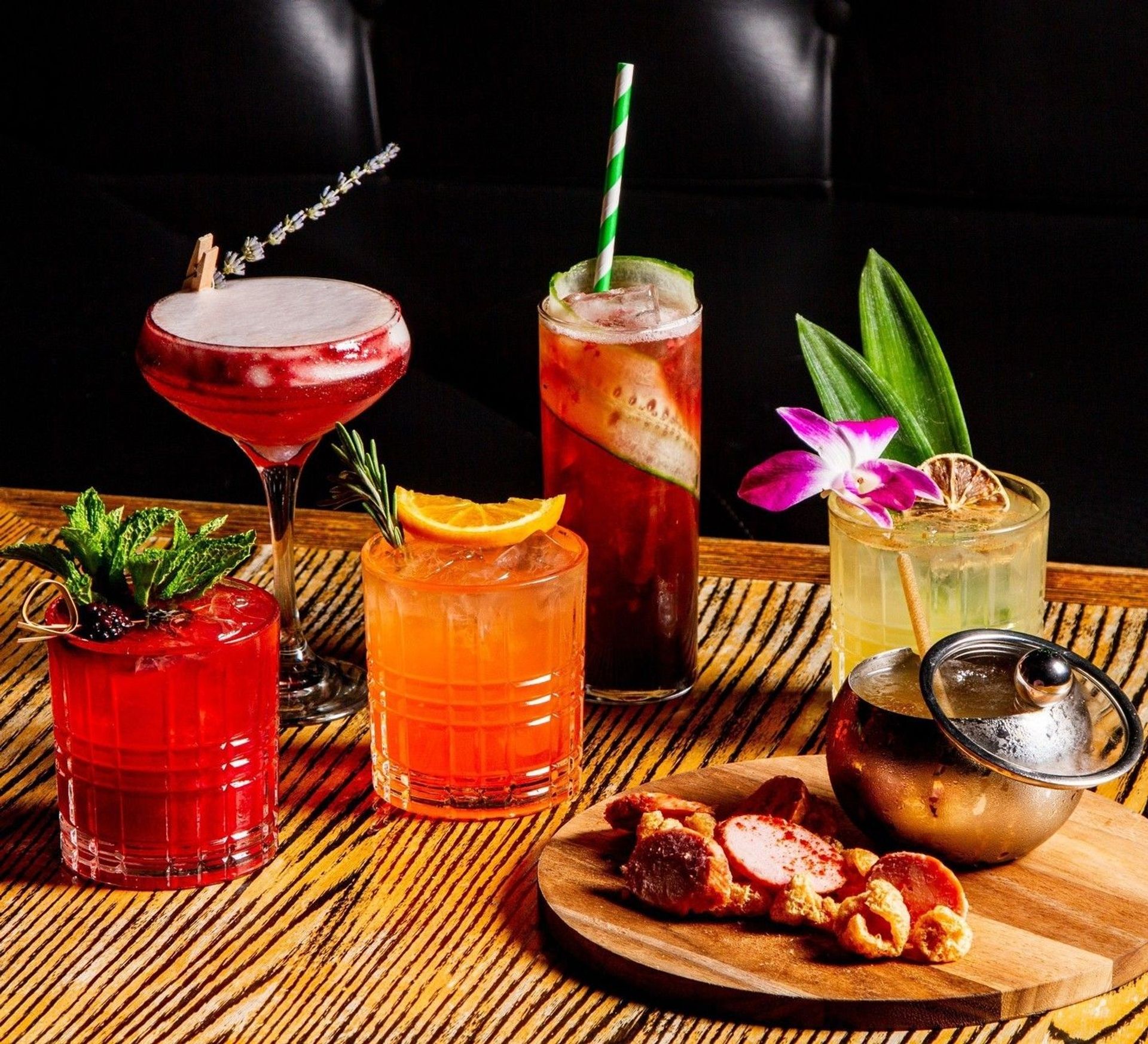 Vibrant craft cocktails on a wooden bar table: frothy red drink with a lavender sprig, mint-garnished ruby cocktail, orange citrus tumbler with rosemary, cucumber-studded highball with a striped straw, orchid-topped tropical cocktail, and a wooden board of charcuterie and crispy snacks.