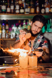 Bartender torching citrus zest over two flaming craft cocktails on a colorful, neon-lit cocktail bar counter