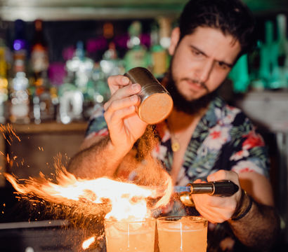 Bartender torching citrus zest over two flaming craft cocktails on a colorful, neon-lit cocktail bar counter