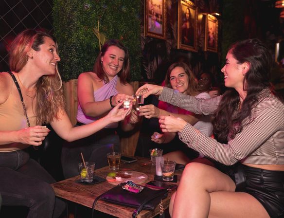 Four women laughing and clinking shot glasses at a cozy urban bar lounge, girls' night out with cocktails on a wooden table