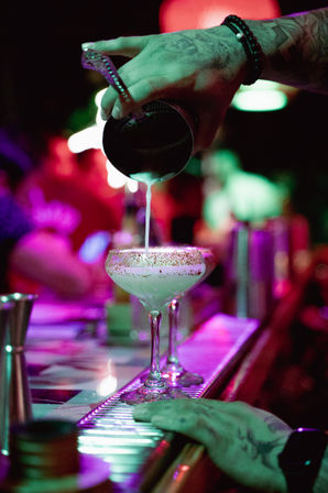 Tattooed bartender pouring a creamy cocktail from a shaker into a sugar-rimmed coupe glass on a neon-lit bar with vibrant purple and green lighting
