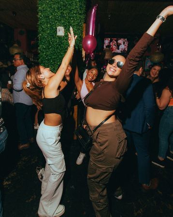 Friends dancing on a crowded indoor nightclub dance floor—two women in front smiling and raising their arms with balloons and a green wall backdrop.
