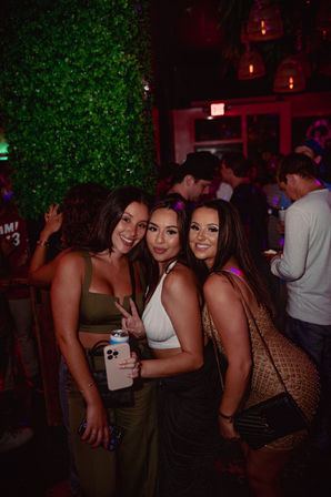 Three smiling women posing at a city nightclub — holding drinks and a phone, neon lighting, faux greenery wall and a crowded bar scene behind them.