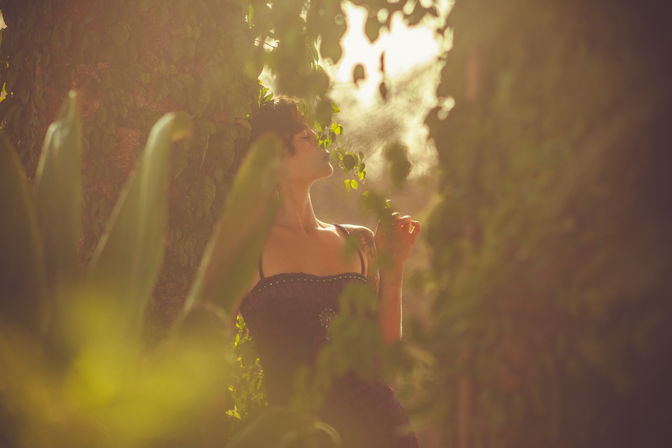 Outdoor portrait of a woman in a strapless vintage dress smelling a vine in a sunlit garden at golden hour, warm tones and soft bokeh