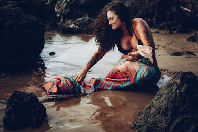 Smiling tattooed woman in a vibrant mermaid tail and black bikini top sits on a rocky beach tide pool, touching her patterned tail and holding a decorative shell.