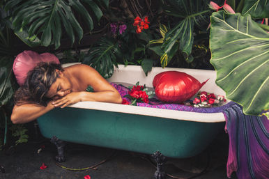 Woman relaxing in a teal clawfoot bathtub outdoors amid lush tropical foliage, a red heart-shaped balloon, and scattered roses on a purple cloth.