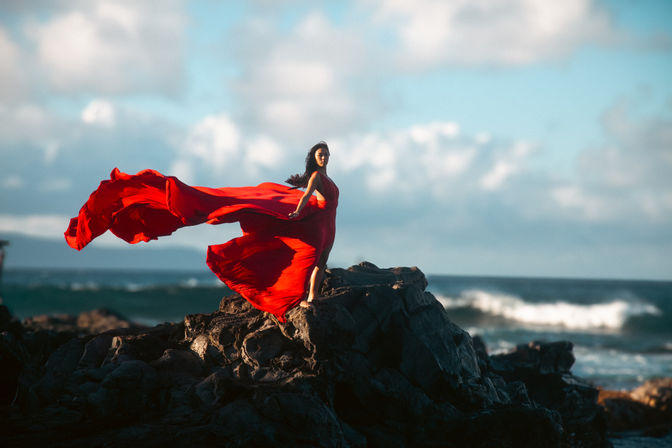 Windswept woman in a flowing red gown standing on dark rocky shoreline, dress billowing dramatically against blue sky and ocean waves.