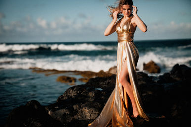 Person in a flowing metallic gold evening gown standing barefoot on dark volcanic rocks along a coastal shoreline, wind-tousled hair and ocean waves breaking behind