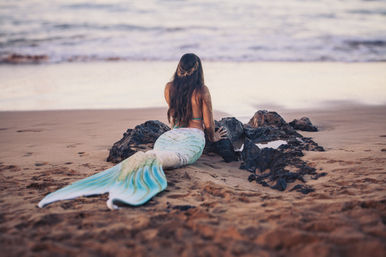 Person with a pastel teal mermaid tail reclining on a sandy beach, facing the ocean and leaning on dark coastal rocks beside a small tide pool.